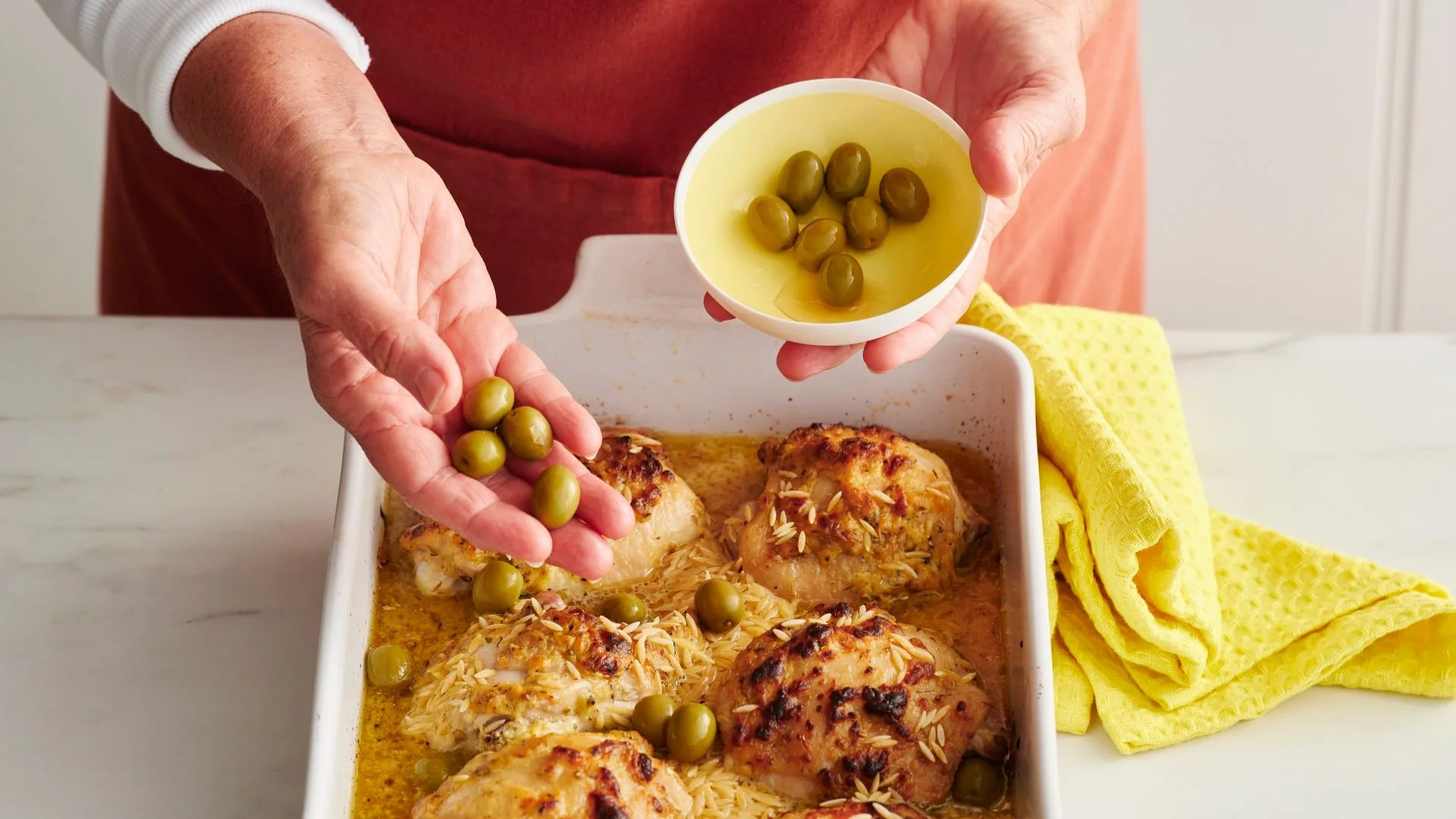 A hand sprinkles olives over cooked lemon chicken thighs in a white pan with orzo