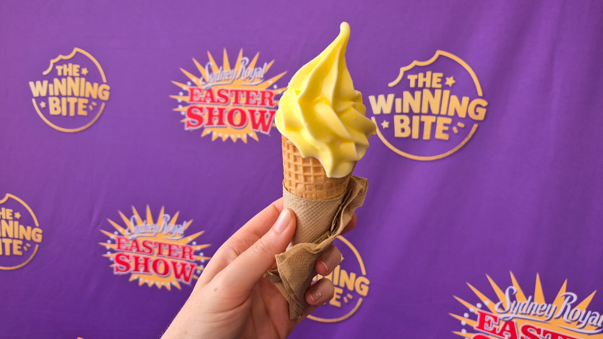 A hand holds an ice-cream cone in front of a purple Sydney Royal Easter Show 2026 banner