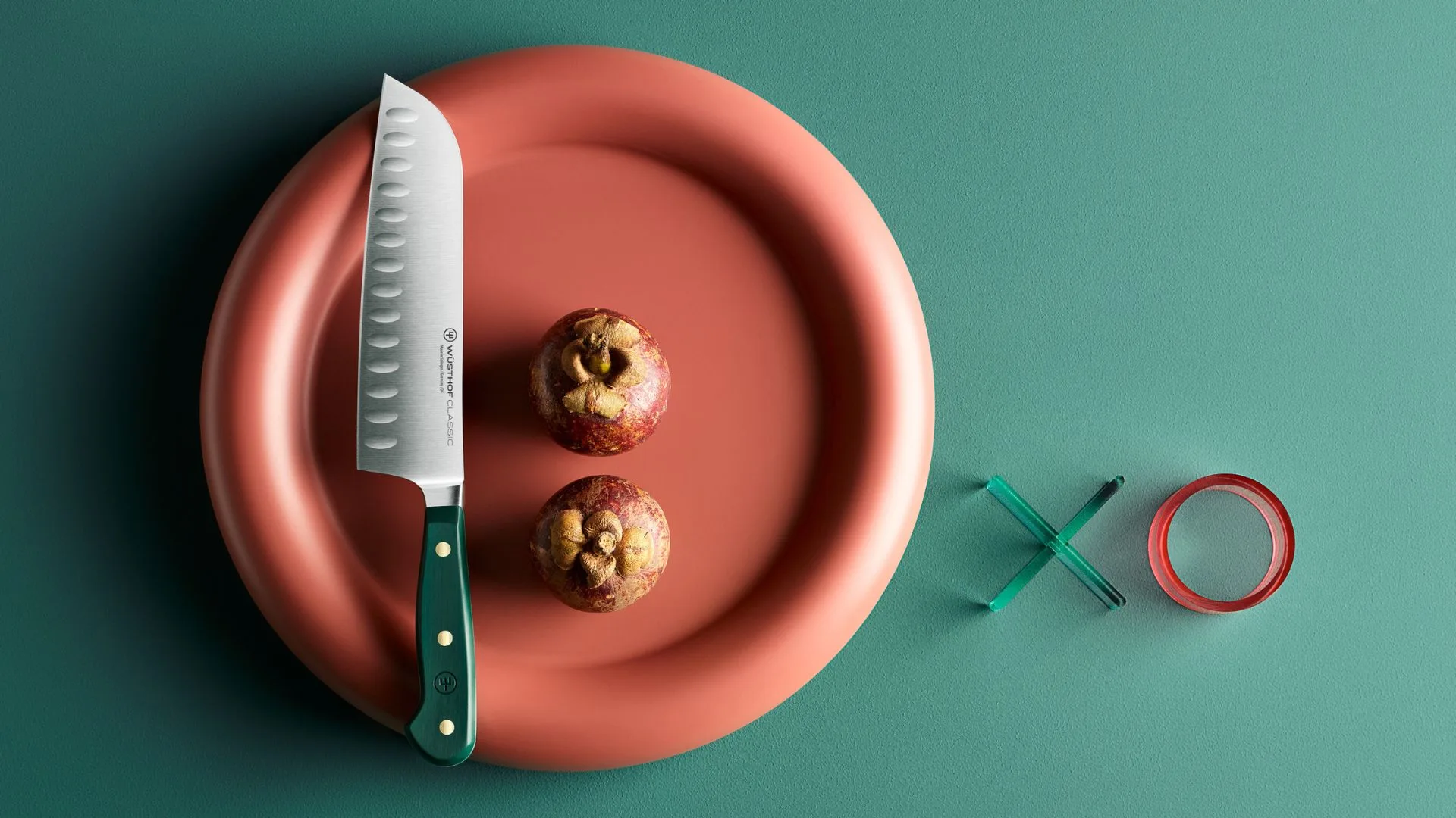 A W&uuml;sthof Classic Santoku knife featuring a "Fresh Rosemary" green handle, resting on a round, salmon-coloured plate. Two mangosteens are positioned next to the scalloped blade. The background is a flat teal surface decorated with transparent "X" and "O" acrylic shapes.