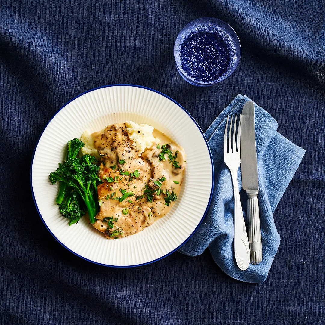 serving of creamy chicken and mushroom on a white plate with a knife and fork beside it and a glass of water