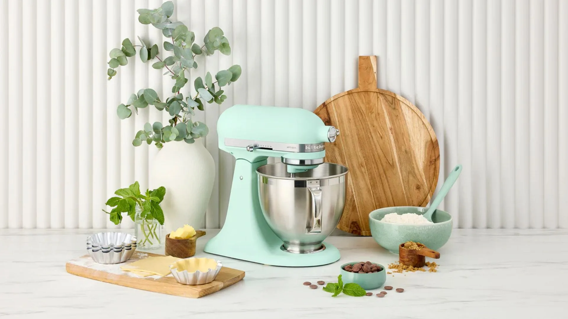 A KitchenAid Artisan stand mixer in Spearmint - KitchenAid's Colour of the Year 2026 - styled on a marble benchtop with baking ingredients including butter, chocolate, flour and fresh mint.