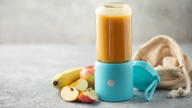 A compact, cordless portable blender with a blue base and clear glass jar containing a blended orange smoothie. It is styled on a textured countertop with fresh apple slices, a whole banana, and a beige drawstring travel bag in the background.