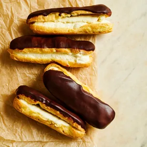 Four long, cream-filled chocolate eclairs on brown baking paper