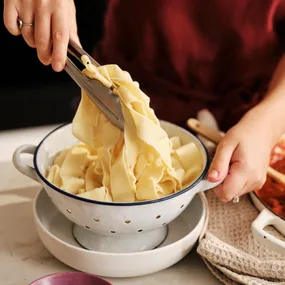 fresh egg pasta dough in a white colander being lifted with tongs