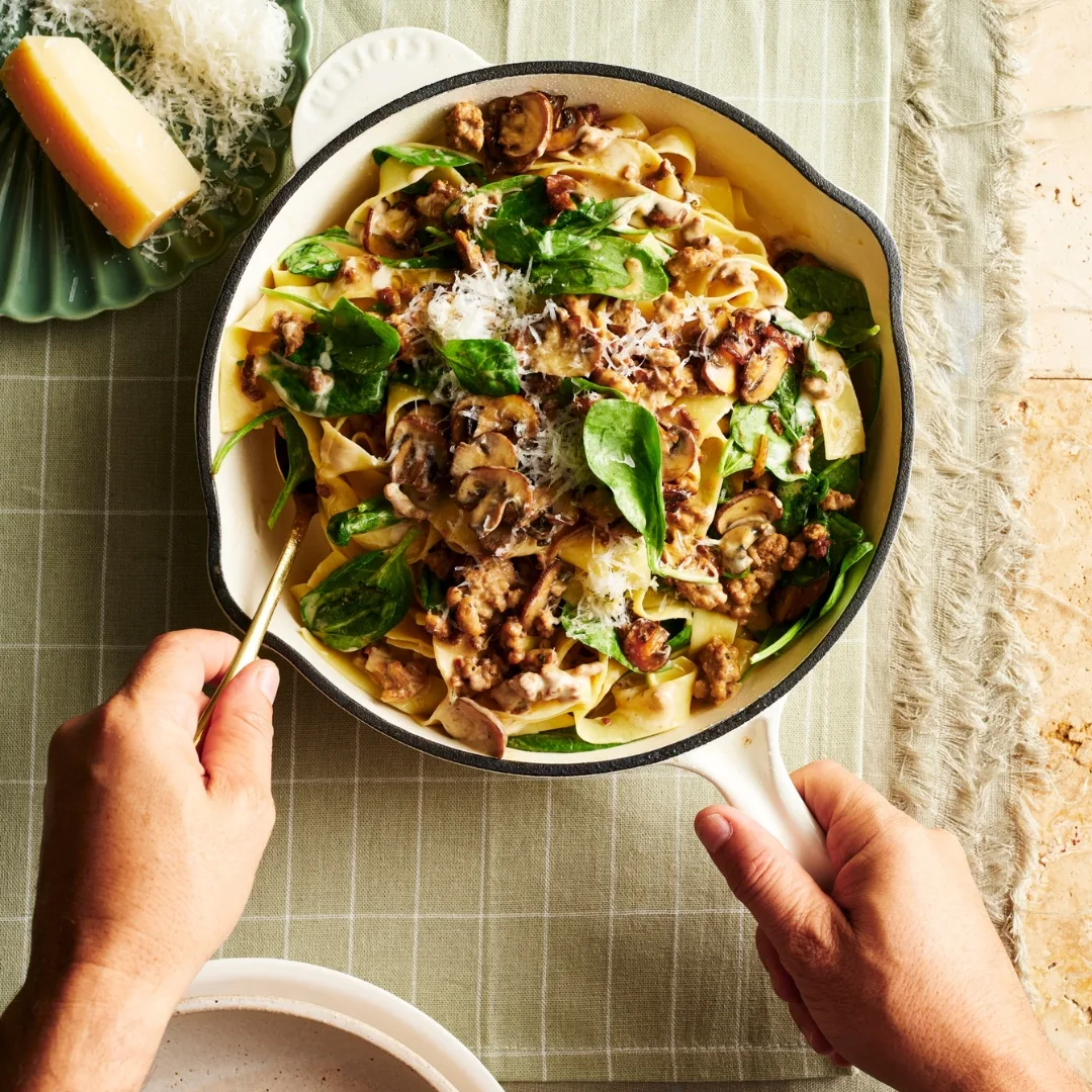 pork mince pasta with spinach and mushroom in a white frying pan with a hand holding a spoon