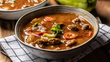 A hearty bowl of Hungarian goulash soup featuring tender beef, potatoes, and red sweet peppers in a rich, paprika-infused broth, garnished with fresh herbs.
