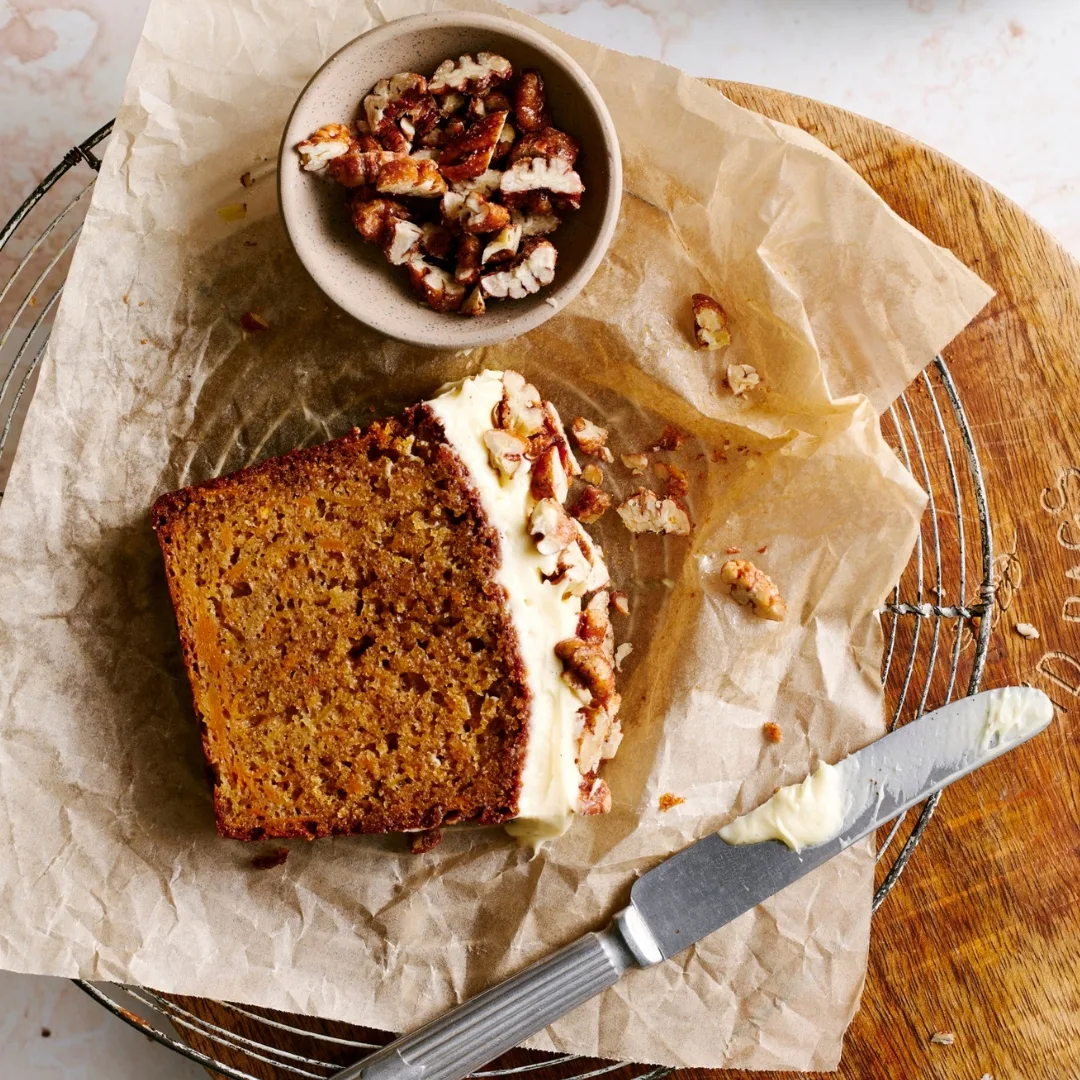 Slice of sweet potato loaf cake on a wooden chopping board with a bowl of pecans