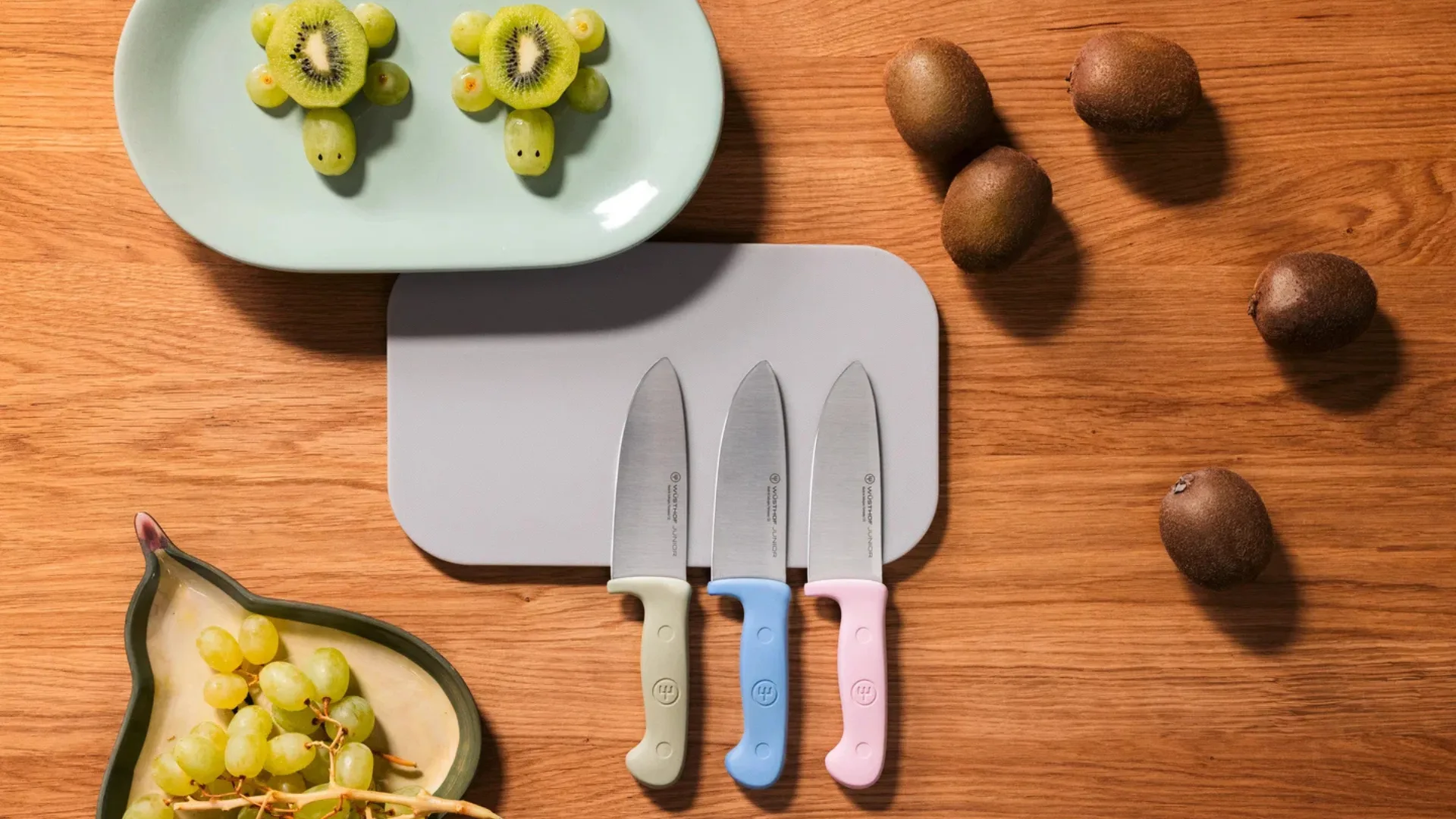 Three Wusthof Junior knives with green, blue, and pink handles resting on a light grey cutting board. They are set on a wooden table alongside whole kiwis, green grapes, and a plate featuring fruit snacks shaped like turtles made from kiwi slices and grapes.
