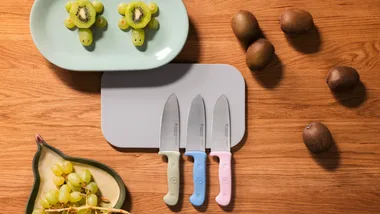 Three Wusthof Junior knives with green, blue, and pink handles resting on a light grey cutting board. They are set on a wooden table alongside whole kiwis, green grapes, and a plate featuring fruit snacks shaped like turtles made from kiwi slices and grapes.
