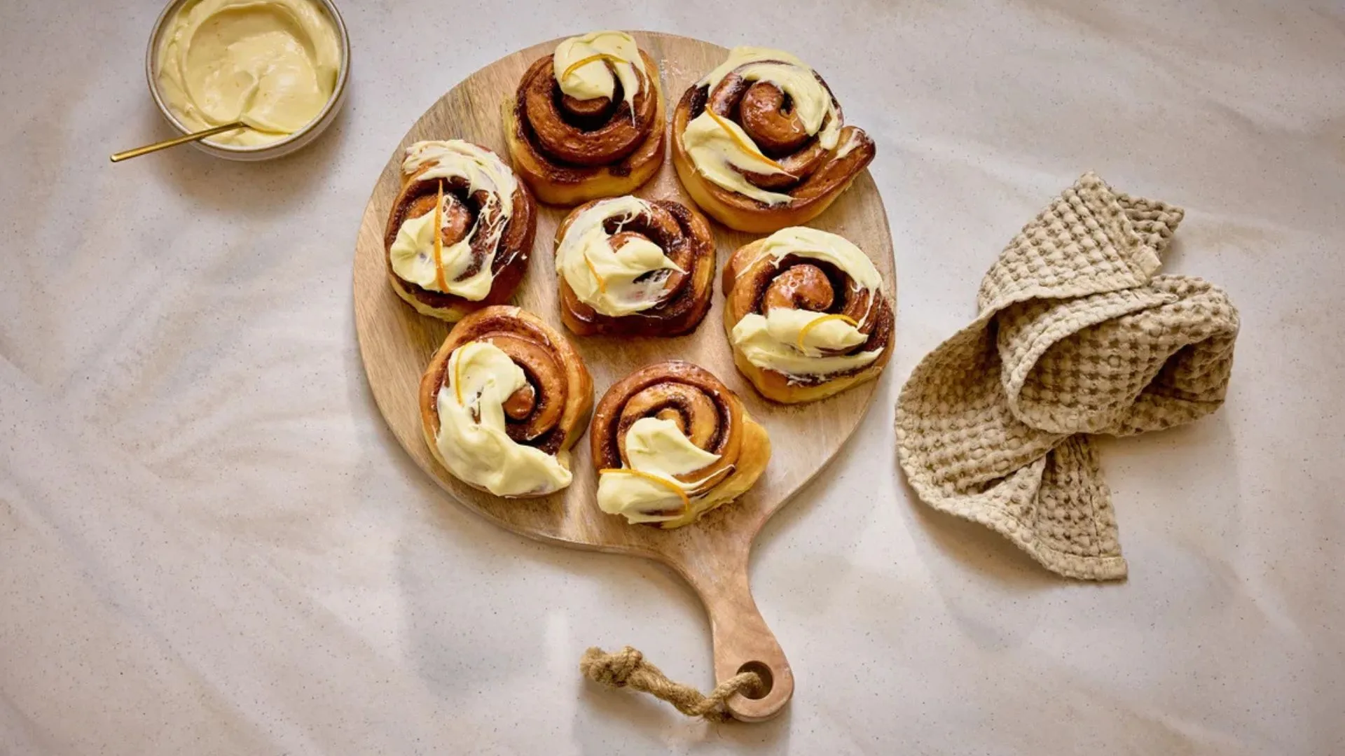 Freshly baked cinnamon and wattleseed fika buns topped with orange zest cream cheese frosting on a wooden board.