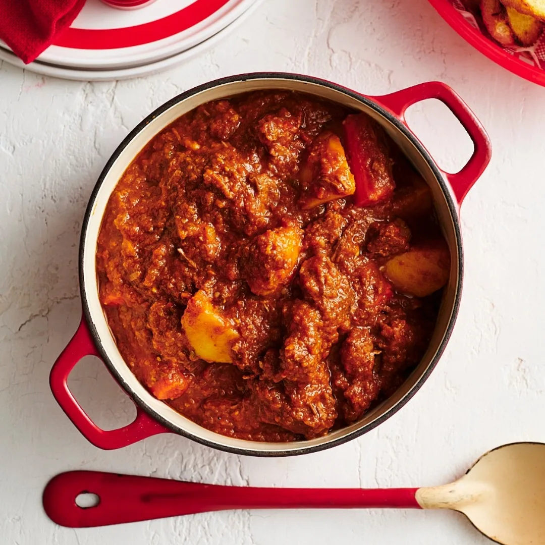 Slow cooker Spanish gravy beef stew in a red saucepan with a metal serving spoon.