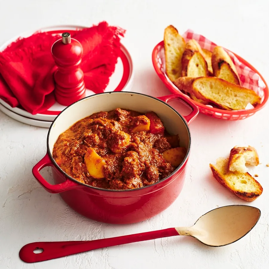 Julie Goodwin's slow cooker beef stew in a red saucepan with a metal serving spoon, and a basket of bread.