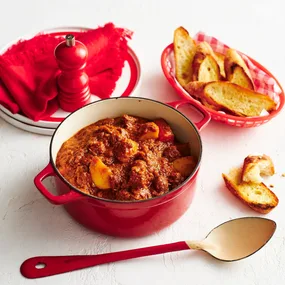 Julie Goodwin's slow cooker beef stew in a red saucepan with a metal serving spoon, and a basket of bread.