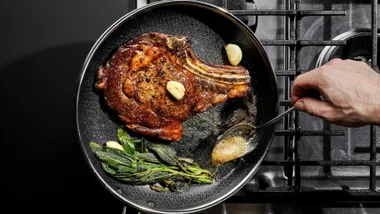 Top-down view of a thick bone-in ribeye steak searing in a HexClad skillet on a gas stovetop, being basted with butter using a spoon, alongside garlic cloves and fresh herbs. Featured in AWWF's article - Hexclad prime day deals.