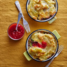 Two individual cottage pies served in small round ceramic baking dishes on a warm golden-brown fabric tablecloth. The dishes have light green handles and dark rims, giving them a rustic, home-cooked presentation.