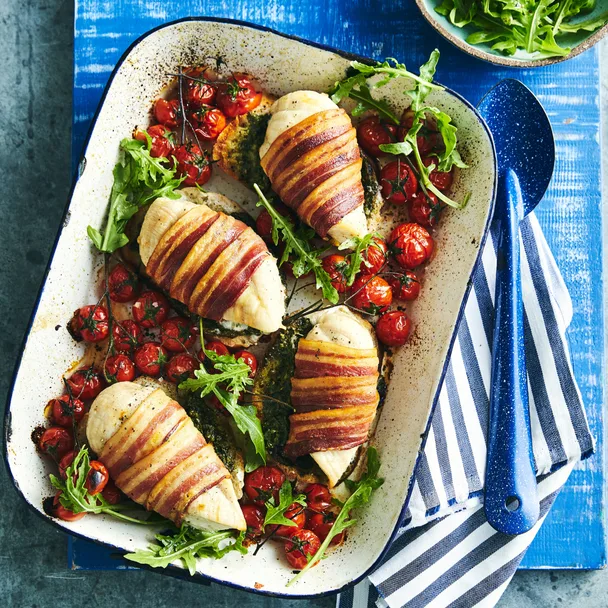 Roasted bacon-wrapped chicken breasts with pesto, cherry tomatoes, and arugula in a baking dish.