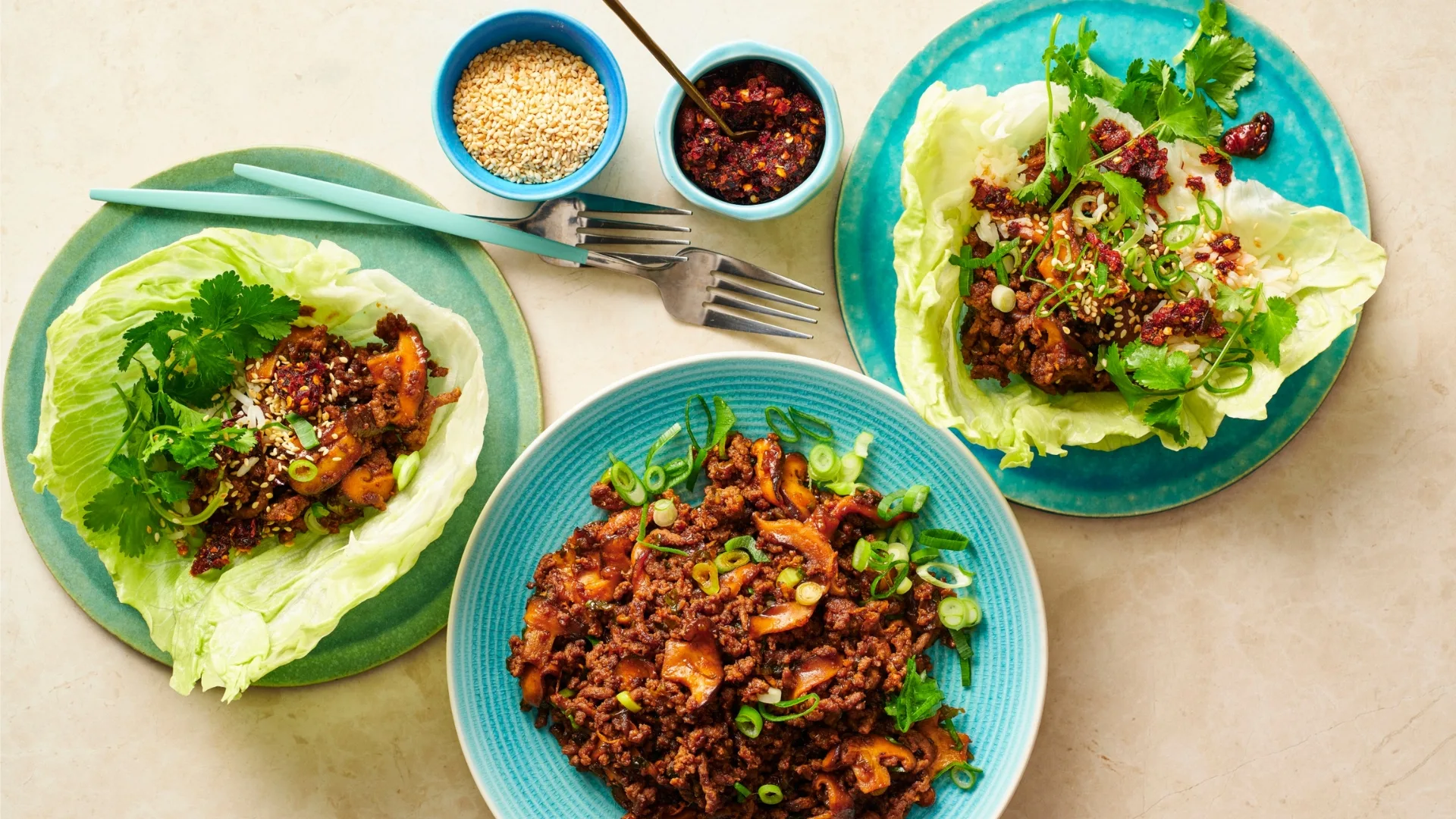 three plates with san choy bow beef lettuce cups, and two small bowls with sesame seeds and chilli paste