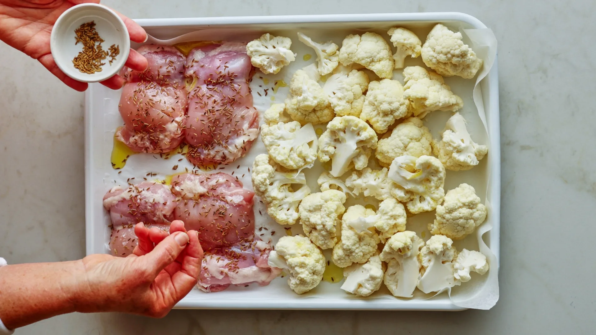 cumin seeds being sprinkled over chicken thigh and cauliflower on a baking tray