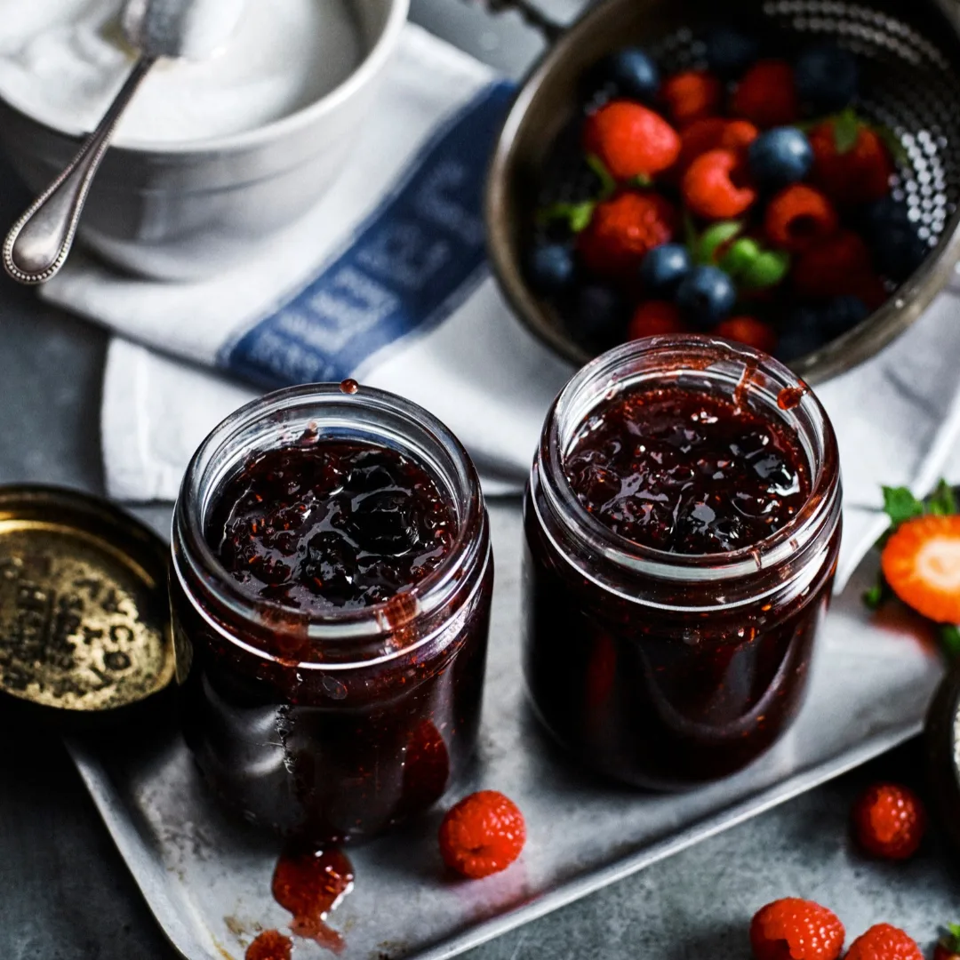 two jars of slow cooker mixed berry jam with a bowl of sugar and strainer of berries