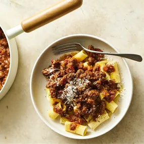 Bowl of rigatoni pasta topped with rich beef Bolognese sauce, grated Parmesan cheese, and cracked black pepper, served on a light stone surface with a fork and saucepan partially visible.