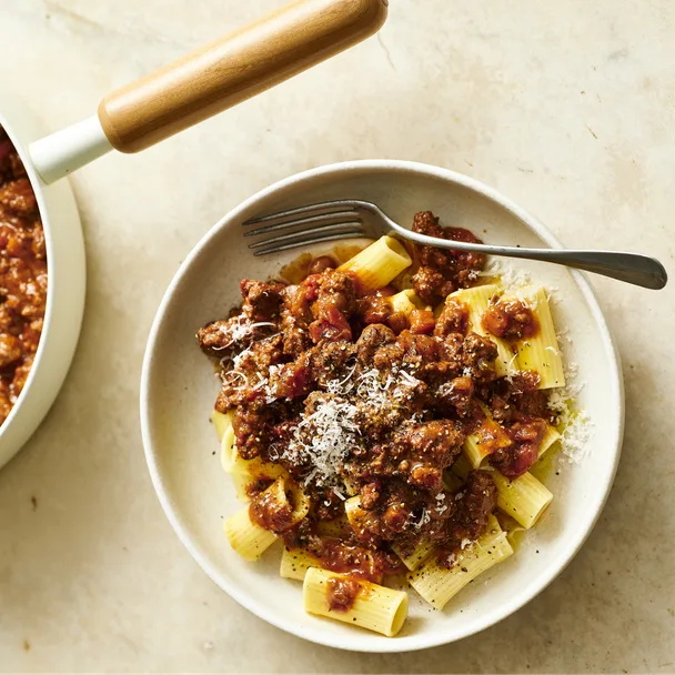 Bowl of rigatoni pasta topped with rich beef Bolognese sauce, grated Parmesan cheese, and cracked black pepper, served on a light stone surface with a fork and saucepan partially visible.