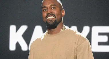 A man in a beige shirt smiles on stage with a blurred "KANYE" text in the background.