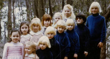 A group of thirteen children with blonde or light-colored hair in the woods, some wearing matching blue outfits.