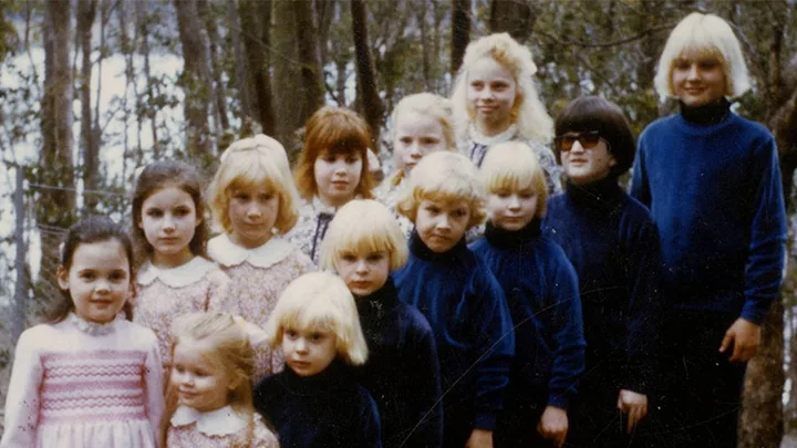 A group of thirteen children with blonde or light-colored hair in the woods, some wearing matching blue outfits.