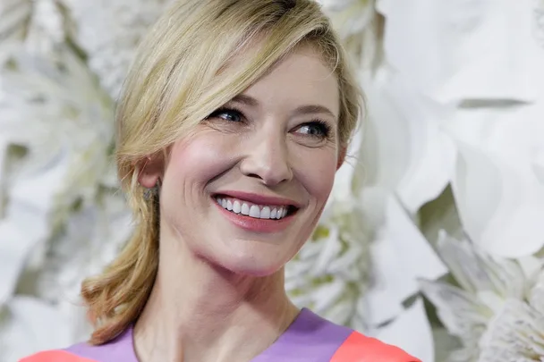 Smiling person with blonde hair in a colorful outfit, standing against a floral background.