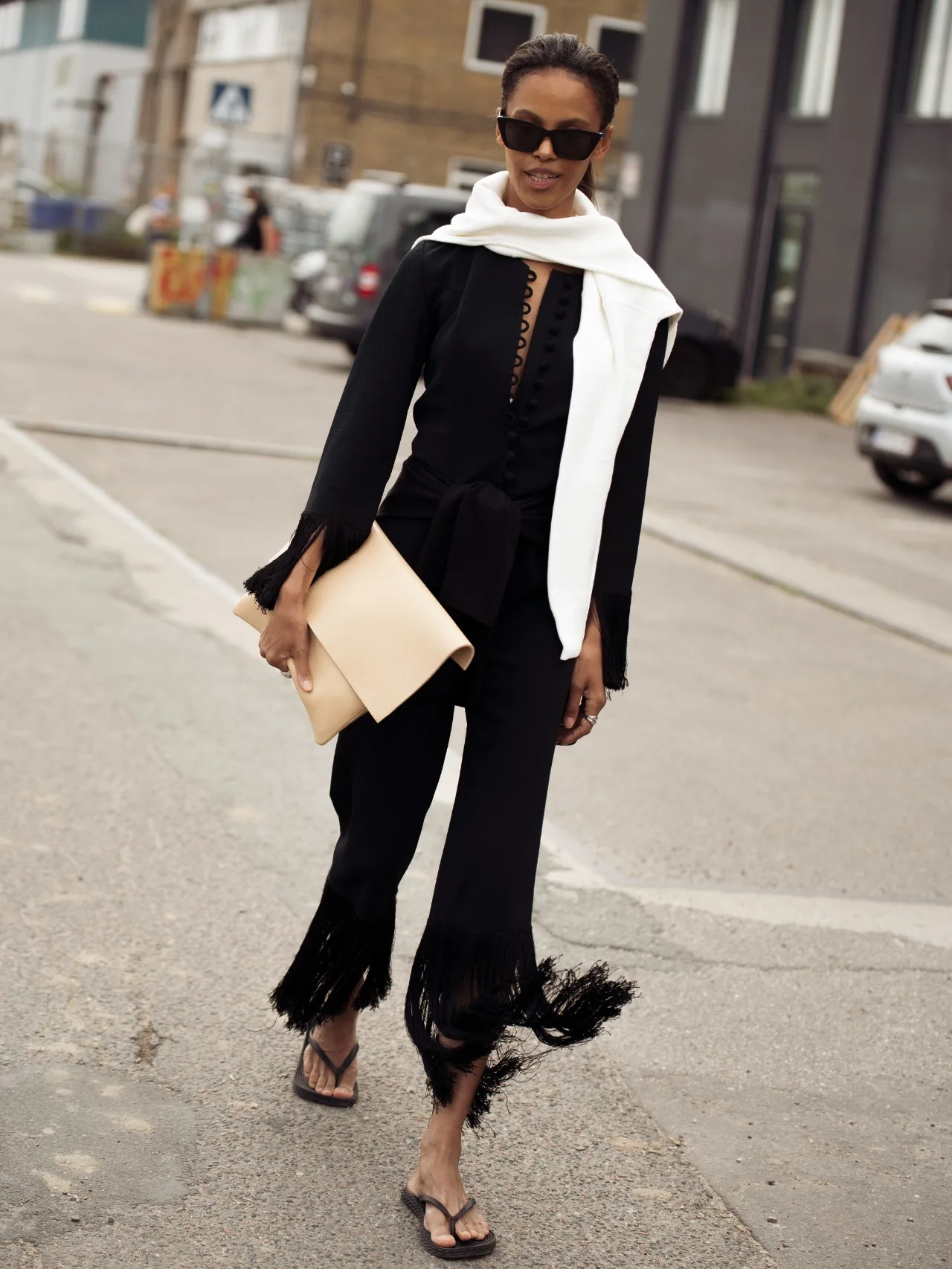 Woman wears all black outfit top and fringed trousers with black thongs, white scarf, black sunglasses and cream oversized clutch bag
