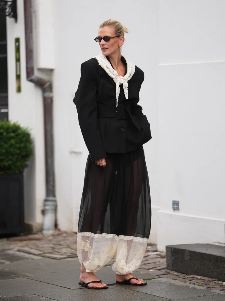 Stylish woman wears intricate sheer black-and-white lace harem trousers with a black blazer and white lace scarf around her neckline with black thongs