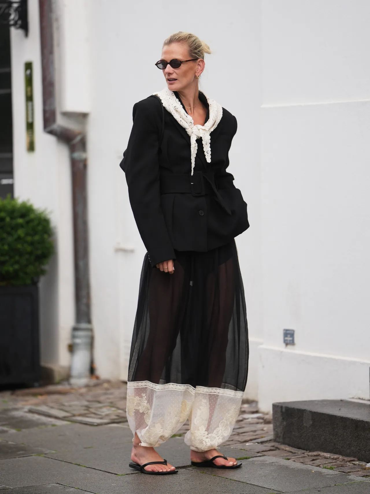 Stylish woman wears intricate sheer black-and-white lace harem trousers with a black blazer and white lace scarf around her neckline with black thongs