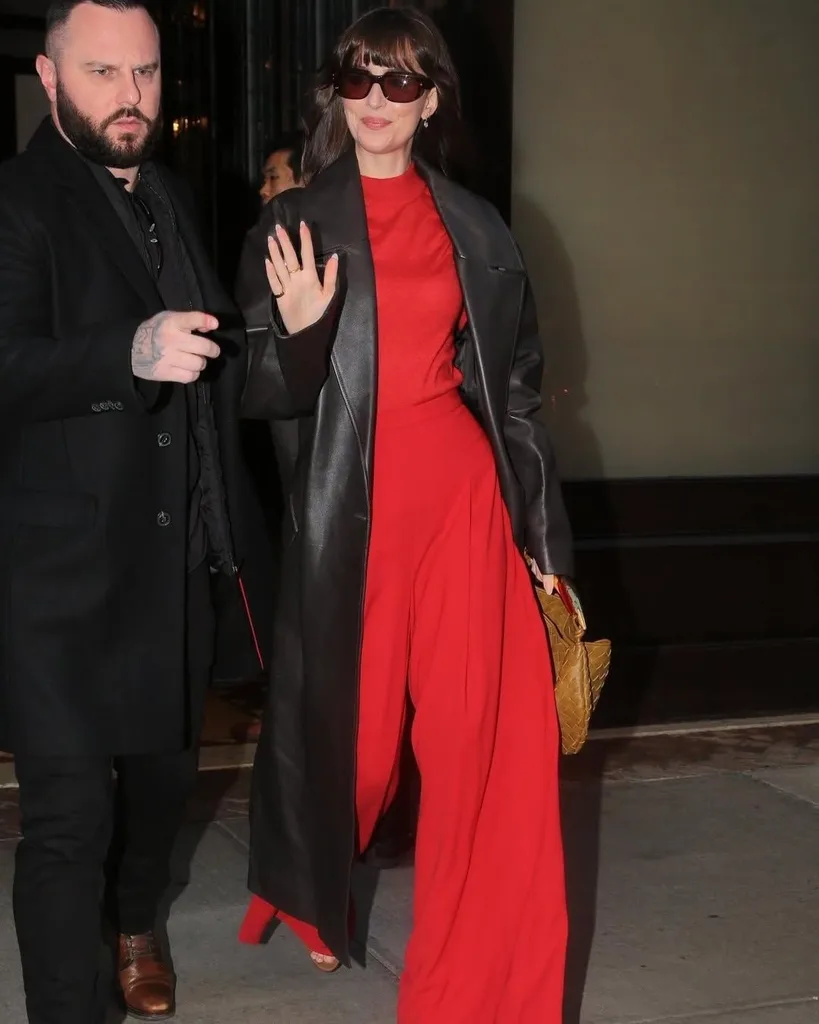 Woman wearing long black leather coat and red jumpsuit waving.