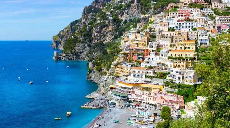 Colorful buildings on a cliff in Positano, Italy, overlook the blue sea with boats and a beach below.