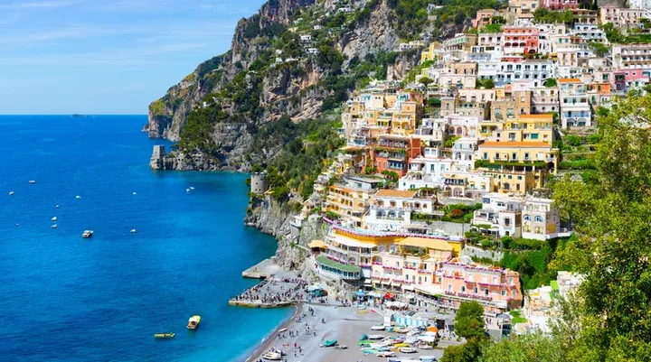 Colorful buildings on a cliff in Positano, Italy, overlook the blue sea with boats and a beach below.