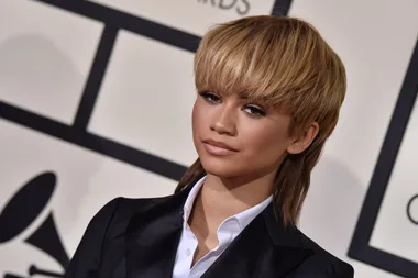 A person with short blonde hair in a black suit at an event with a background featuring a Grammy Award logo.