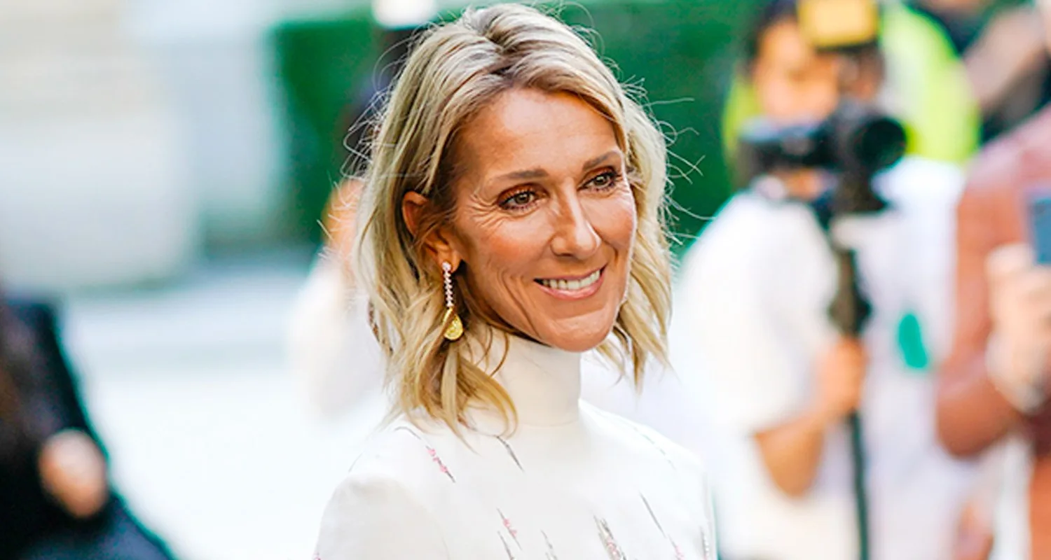 A woman with blonde hair smiles, wearing gold earrings and a white top, with blurred people in the background.
