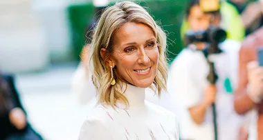 A woman with blonde hair smiles, wearing gold earrings and a white top, with blurred people in the background.