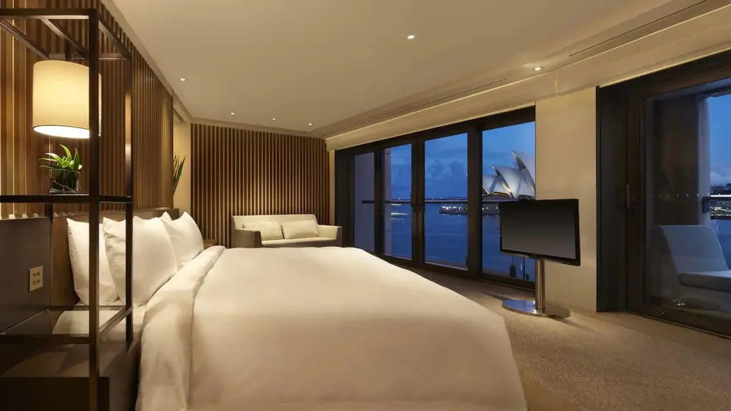 A suite at the Park Hyatt Sydney hotel that looks out to the Sydney Opera House. The room is decorated with neutral colours and a simple timber wood detail on the wall.