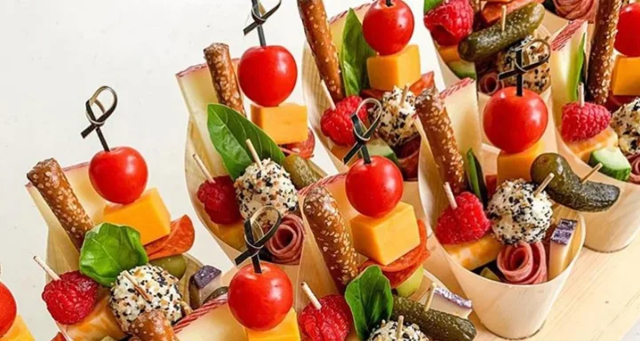 Mini charcuterie cups with cherry tomatoes, cheese cubes, pretzels, cucumber slices, pickles, raspberries, and skewers of seasoned cheese.