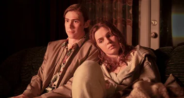 Two young adults sitting closely on a sofa in dim light, one rests their head on the other's shoulder, looking thoughtful.