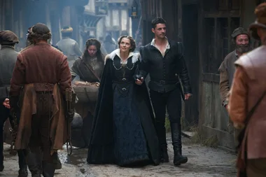 Two characters in period costumes walk through a bustling medieval marketplace.