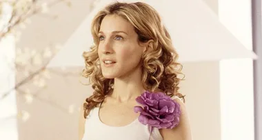 A woman with curly hair, wearing a white top with a large purple flower accessory.