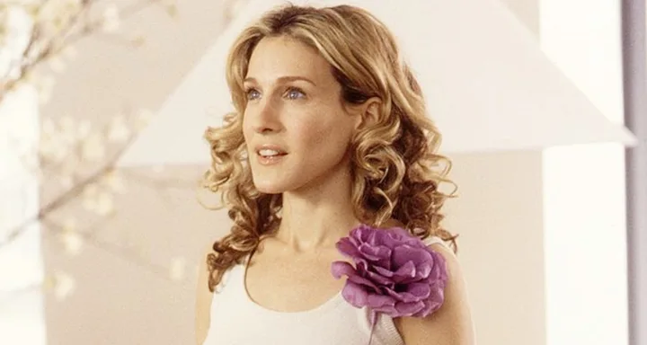 A woman with curly hair, wearing a white top with a large purple flower accessory.