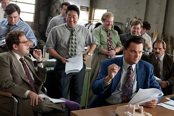 Group of men in an office, one seated in a blue suit holding papers, others standing and smiling, lively discussion.