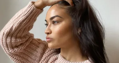 Woman in a pink knit sweater looking thoughtfully to the side, with hand in her hair against a plain background.