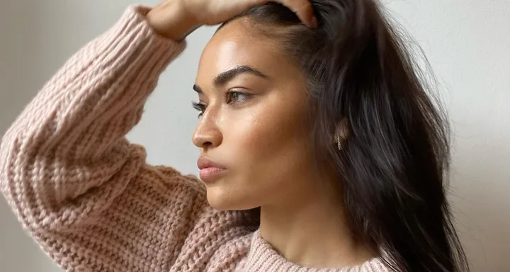 Woman in a pink knit sweater looking thoughtfully to the side, with hand in her hair against a plain background.