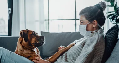 A woman wearing a mask sitting on a couch, petting her dog.