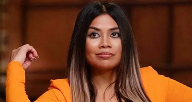A woman with long dark hair and an orange outfit, sitting confidently with a raised hand in a brown room.