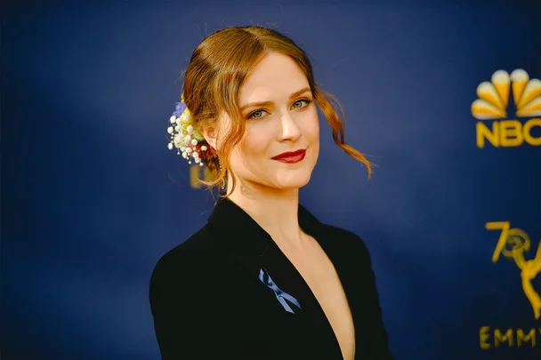 A person with red lipstick and floral hair accessory, wearing a black outfit, at an event with NBC and Emmy logos.